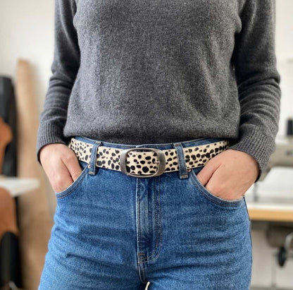 Gros plan sur la taille d'une femme portant la ceinture artisanale Uncia Labossa, en cuir poil naturel imprimé panthère des neiges, avec un jean bleu et un pull gris. Ses mains sont dans les poches. La boucle argentée brossée est bien visible.